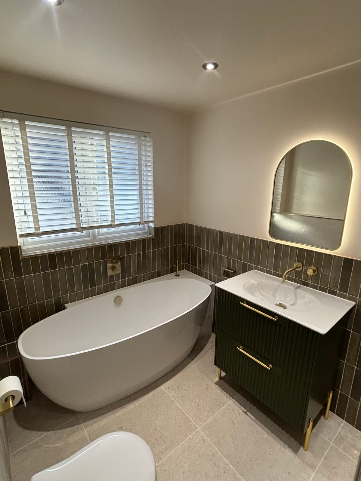 Freestanding bath beside a green-tiled wall with a fluted vanity unit and brass taps